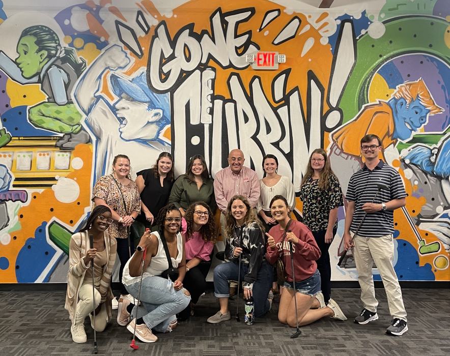 A dozen people stand in front of a graffitied wall, smiling and posing with mini golf clubs. 