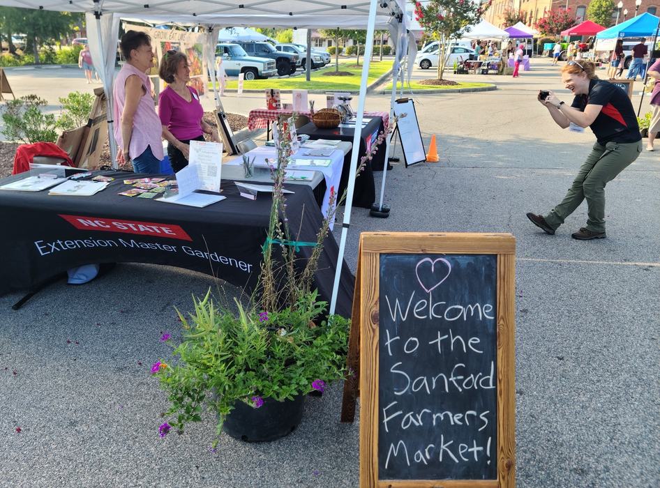 Two volunteers smile while someone takes a picture of their information tent at the Sanford Farmers Market.