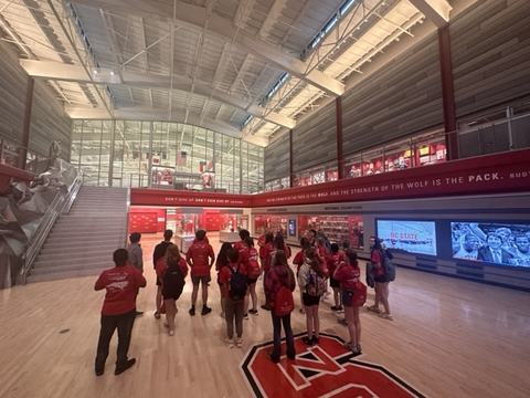 Students in red shirts gathered in NC State arena lobby with large "S" logo on floor