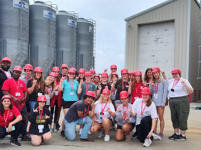 Group of people wearing red hard hats posing and making hand signs in front of metal grain silos
