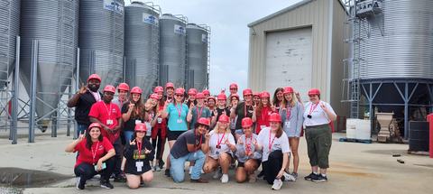 Group of people wearing red hard hats posing and making hand signs in front of metal grain silos