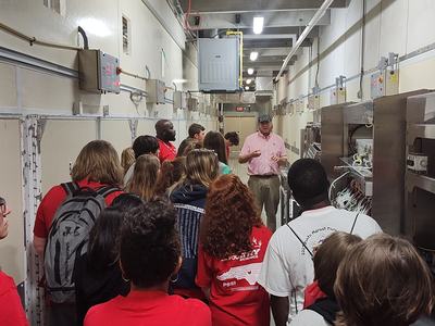 Group touring an industrial facility, guide speaking beside large machinery