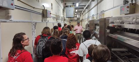 Group touring an industrial facility, guide speaking beside large machinery