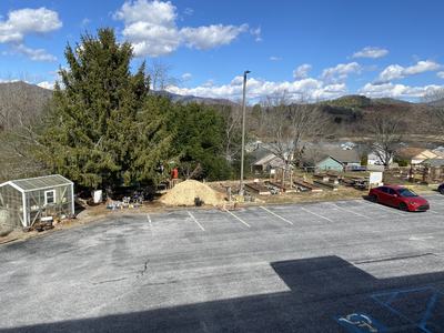 Parking lot with one red car, greenhouse, large evergreen, mulch pile and raised garden beds