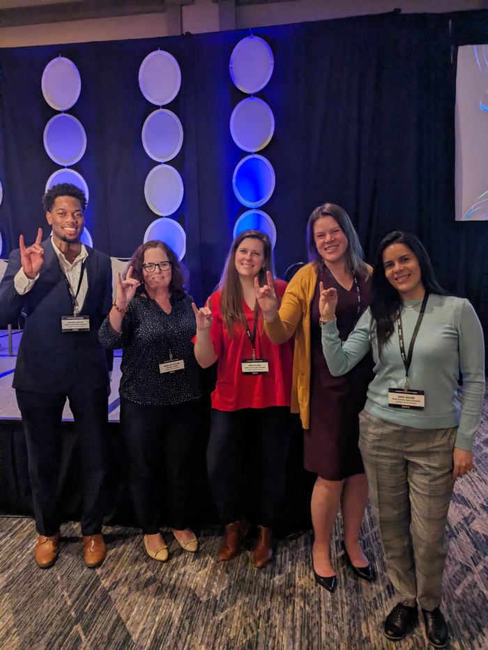 Participants in front of conference background, hold up wolfpack hand sign. 