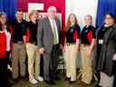 Seven people standing in a row at an indoor event; five wear black shirts and beige pants.