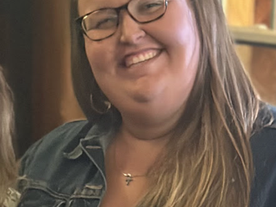 Adult woman with glasses and long hair wearing a denim jacket and cross necklace, smiling