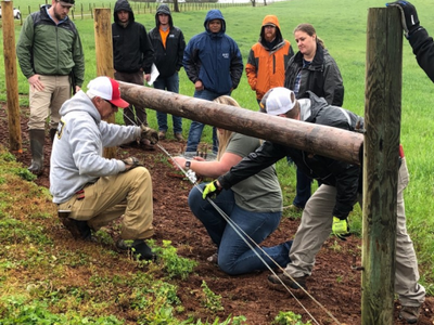 Several people kneeling and standing securing wire to a horizontal wooden fence post in a grassy field