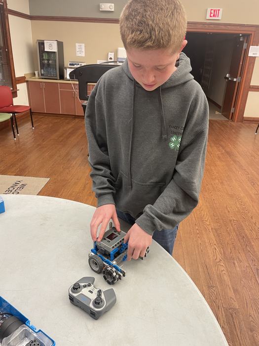 Teen adjusting a small blue-gray robot on a table, controller and EXIT sign visible