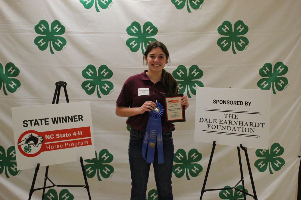 Person holding blue ribbon and plaque in front of 4‑H backdrop and signs: STATE WINNER; THE DALE EARNHARDT FOUNDATION