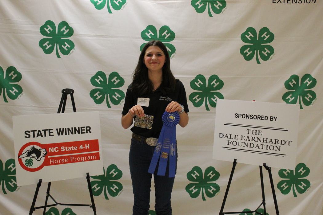 Young woman holding blue ribbon and belt buckle in front of 4‑H backdrop and "STATE WINNER" sign
