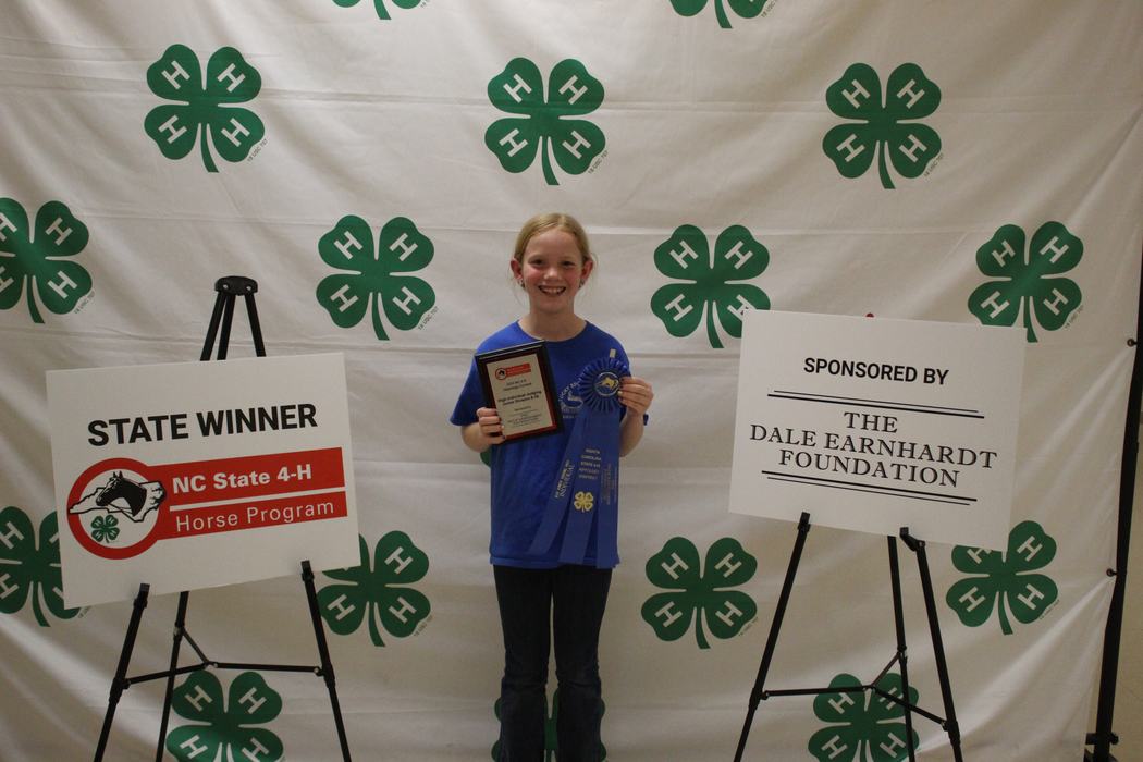 Child holding plaque and blue ribbon in front of 4‑H backdrop; sign reads "STATE WINNER"