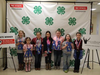 Seven children holding blue ribbons before a 4‑H backdrop and a sign reading STATE WINNER