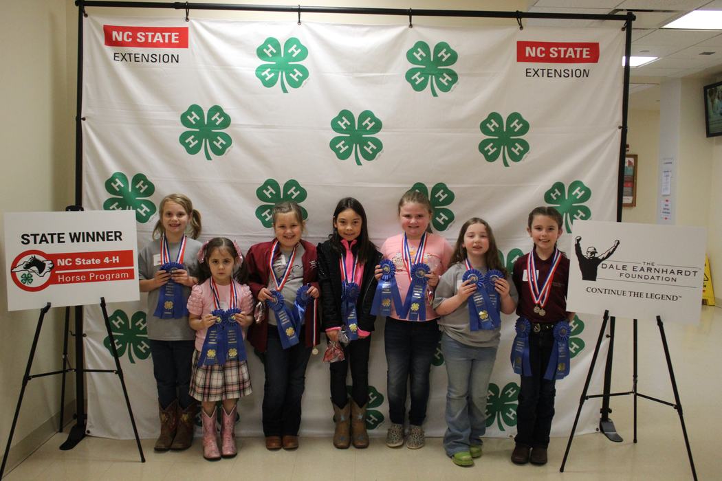 Seven children holding blue ribbons before a 4‑H backdrop and a sign reading STATE WINNER