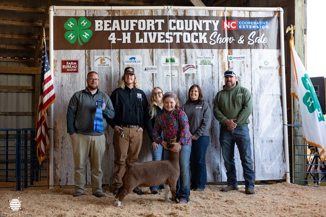 A group of kids and adults posing with a ribbon and a goat.