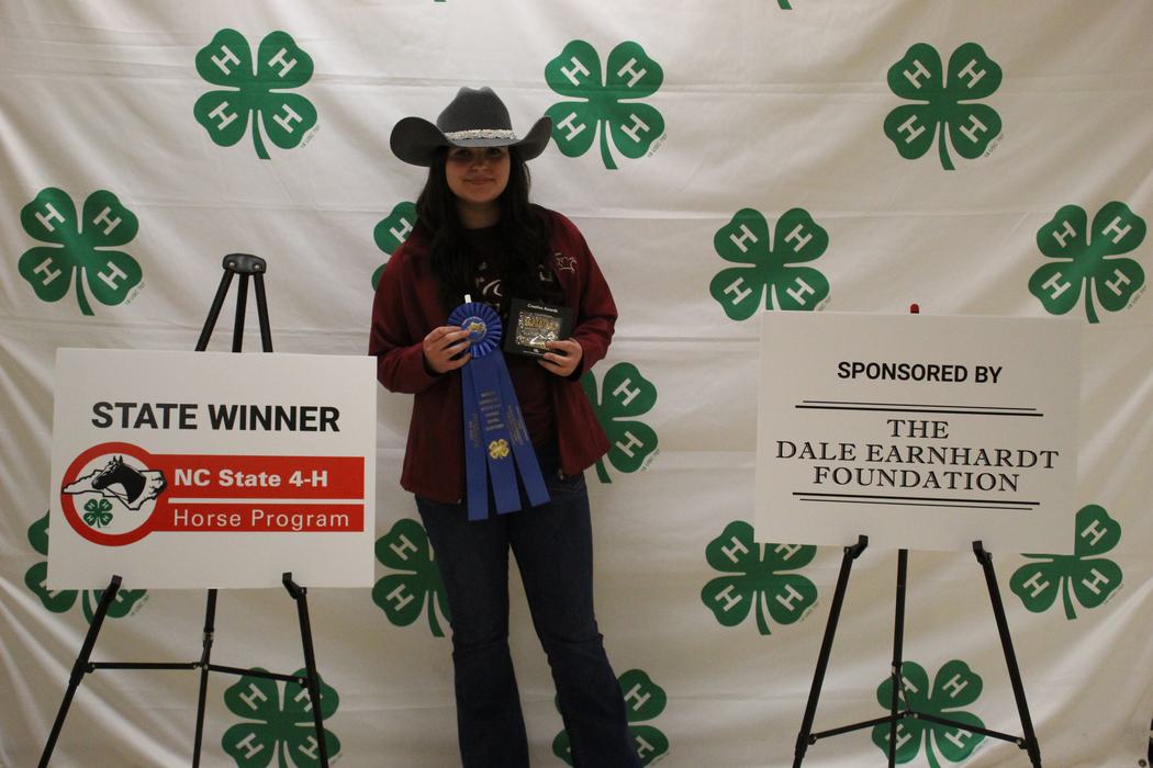 Person in cowboy hat holding blue ribbon and plaque in front of 4-H "STATE WINNER" backdrop