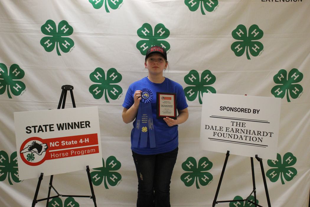 Youth holding blue ribbon and plaque in front of 4‑H backdrop; sign reads "STATE WINNER"