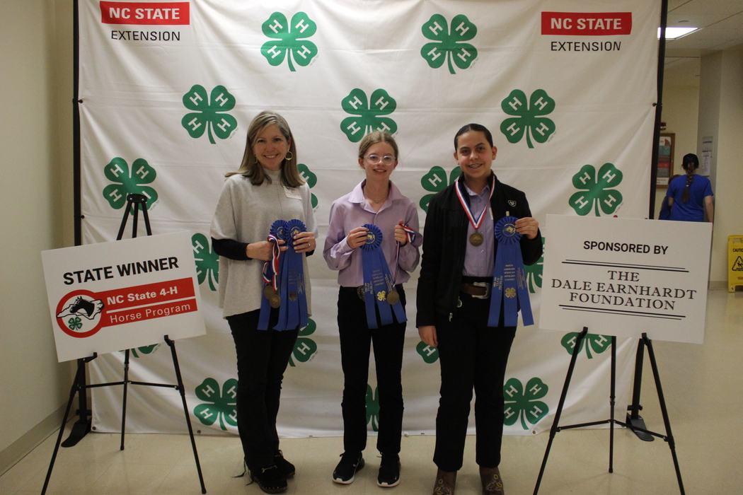 Three people holding blue ribbons before NC State Extension 4-H backdrop; sign "STATE WINNER"