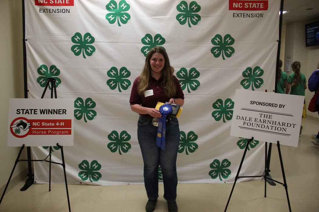 Woman holding blue ribbon and plaque with "STATE WINNER" in front of NC State 4‑H backdrop