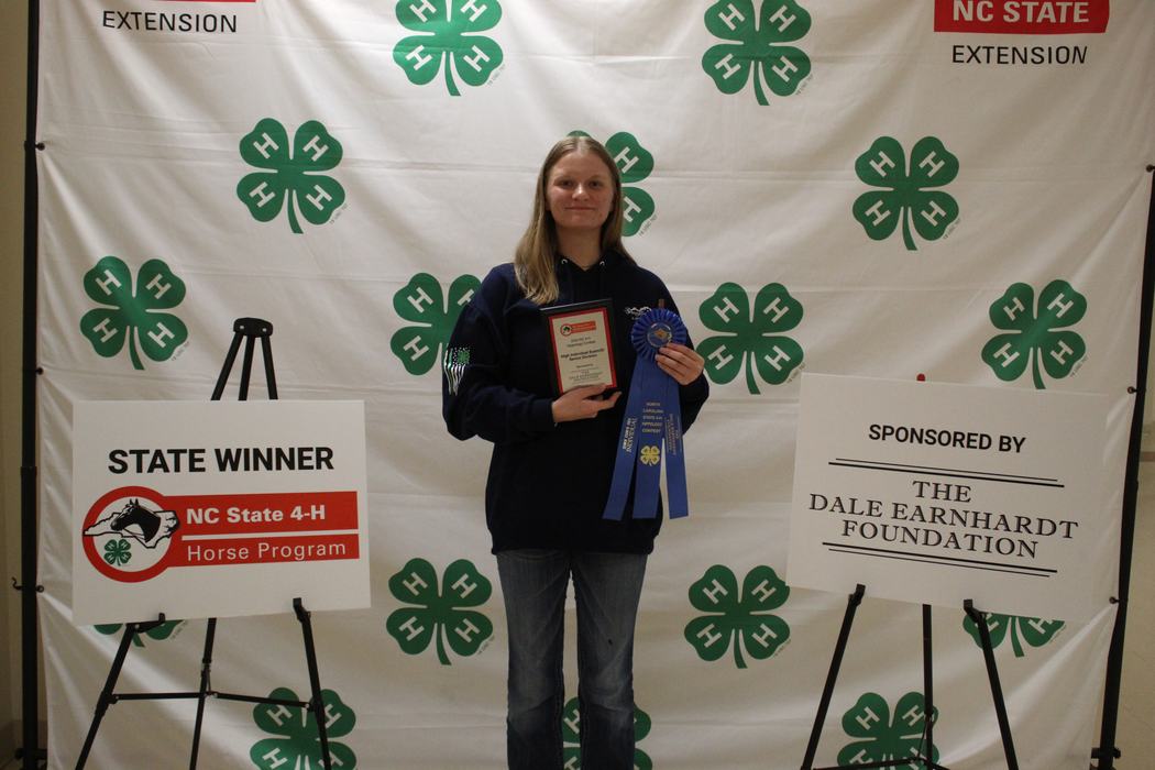 State winner holding plaque and blue ribbon in NC State 4‑H backdrop