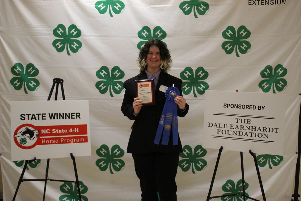 Person holding plaque and blue ribbon in front of 4-H backdrop; sign reads STATE WINNER