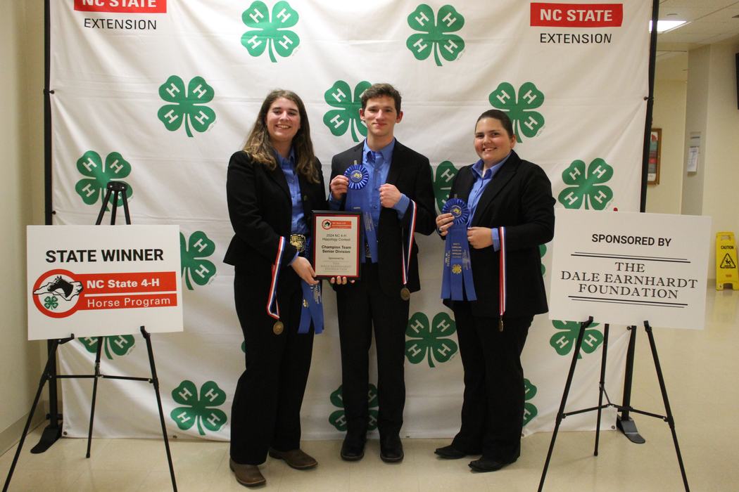 Three people holding ribbons and plaque before NC State 4‑H backdrop; sign reads STATE WINNER
