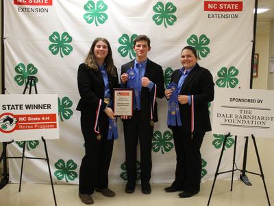 Three people holding ribbons and plaque before NC State 4‑H backdrop; sign reads STATE WINNER