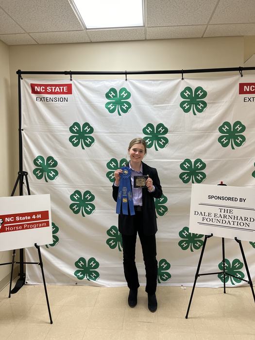 Person holding blue ribbon and plaque before 4‑H backdrop; sign reads "NC State 4‑H Horse Program"