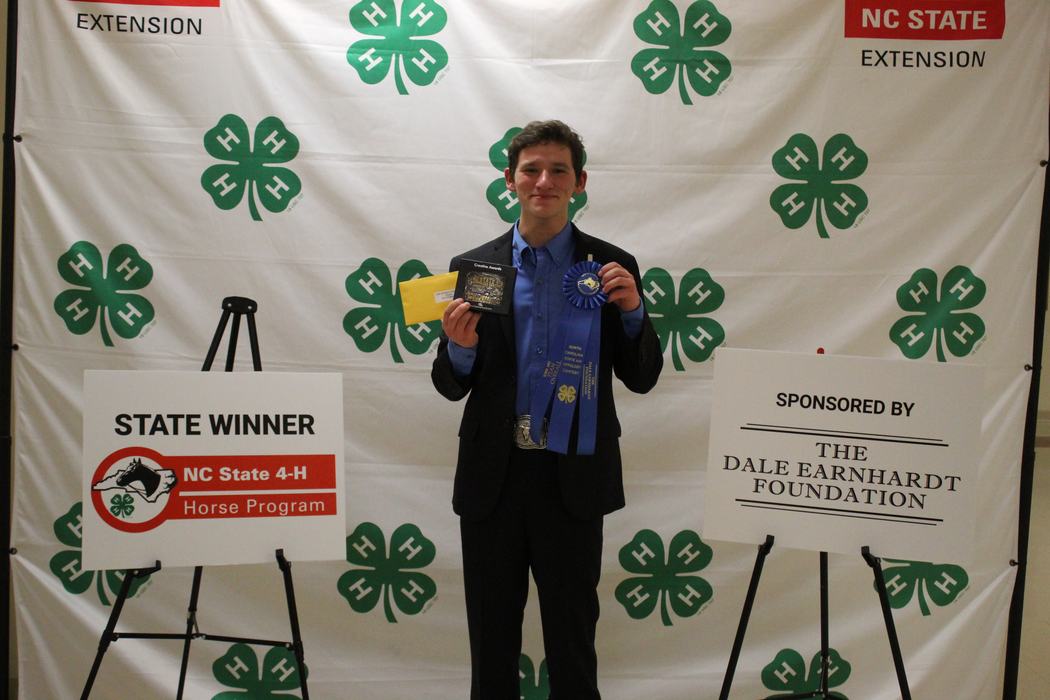 Young man holding blue ribbon and plaque in front of 4‑H backdrop; sign reads STATE WINNER