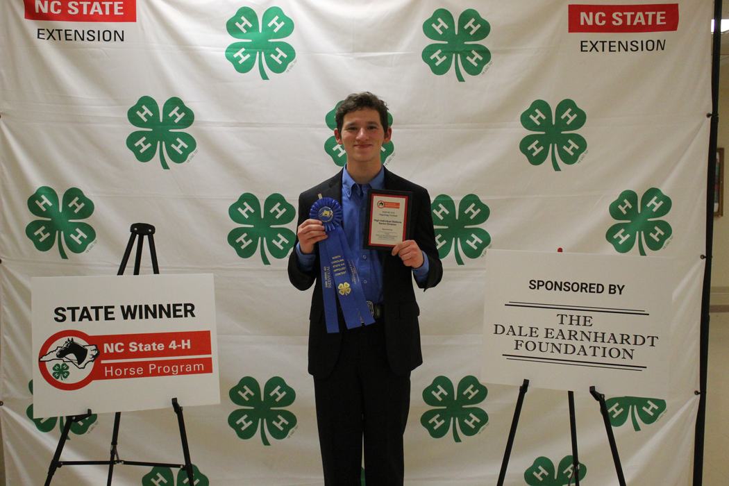 Young man holding blue ribbon and plaque in front of 4‑H backdrop; sign reads "STATE WINNER"