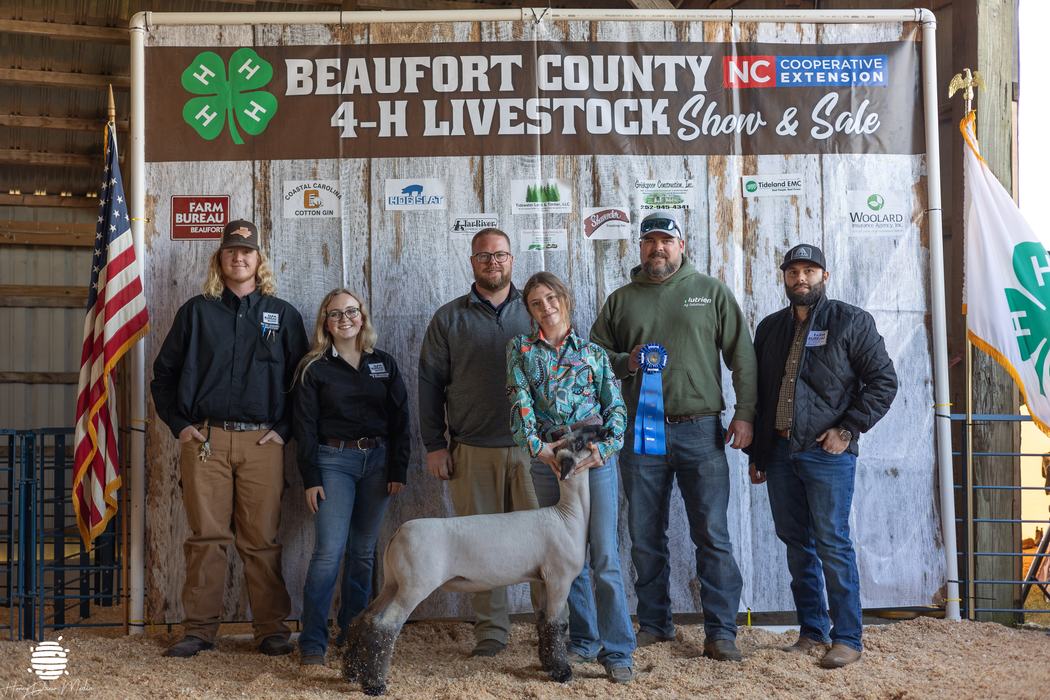 A group of kids and adults posing with a ribbon and a lamb.