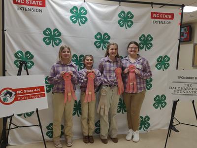 Four girls in matching shirts holding ribbons in front of a 4-H backdrop reading "NC State Extension"
