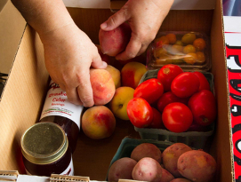 Hands placing peaches into a box labeled "got to be NC AGRICULTURE" with tomatoes, potatoes, jars