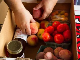 Hands placing peaches into a box labeled "got to be NC AGRICULTURE" with tomatoes, potatoes, jars