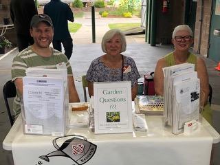 Three people seated at an information table with pamphlets and a "Garden Questions ???" sign.