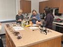 Three women preparing food on a kitchen island with bowls, ingredients, and appliances