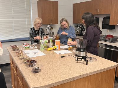Three women preparing food on a kitchen island with bowls, ingredients, and appliances