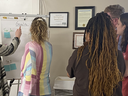 A person writes on a large notepad with four people engaging from the right of the writer. 