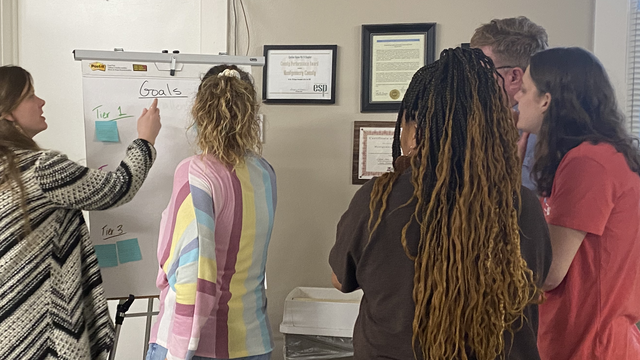 A person writes on a large notepad with four people engaging from the right of the writer. 