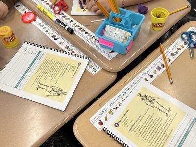 Hands at classroom desks with skeleton worksheets, pencils, Play‑Doh, scissors, water bottle