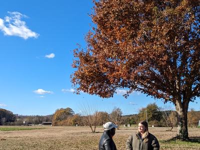 Two people standing and talking under a large autumn tree in an open field