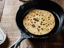 Round flatbread cooking in cast-iron skillet on wood table; watermark 'Anshu A on Unsplash'