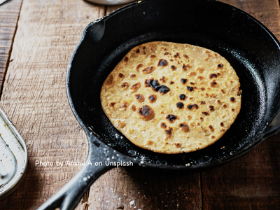 Round flatbread cooking in cast-iron skillet on wood table; watermark 'Anshu A on Unsplash'