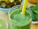 Tall glass of green spinach smoothie with yellow straw, pineapple chunks and lime nearby