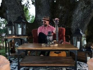 Man wearing hat sitting on red armchair holding guitar upright beside wooden table outdoors