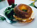 Toasted bagel sandwich with egg and ham on a white plate, arugula salad and fork