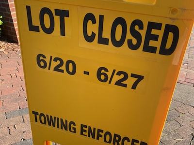 Yellow sign reading: LOT CLOSED 6/20-6/27; TOWING ENFORCED on brick sidewalk