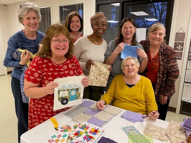 Seven women gathered at a table holding fabric pieces and quilt blocks for quilting.