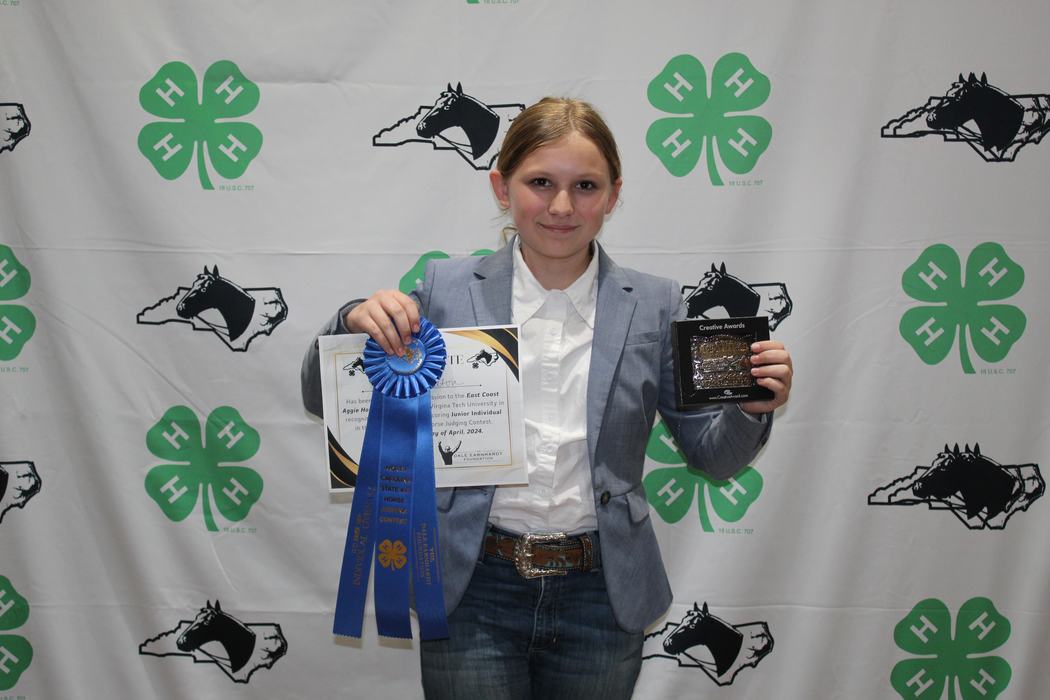 Young person holding blue ribbon, certificate and plaque in front of 4‑H backdrop
