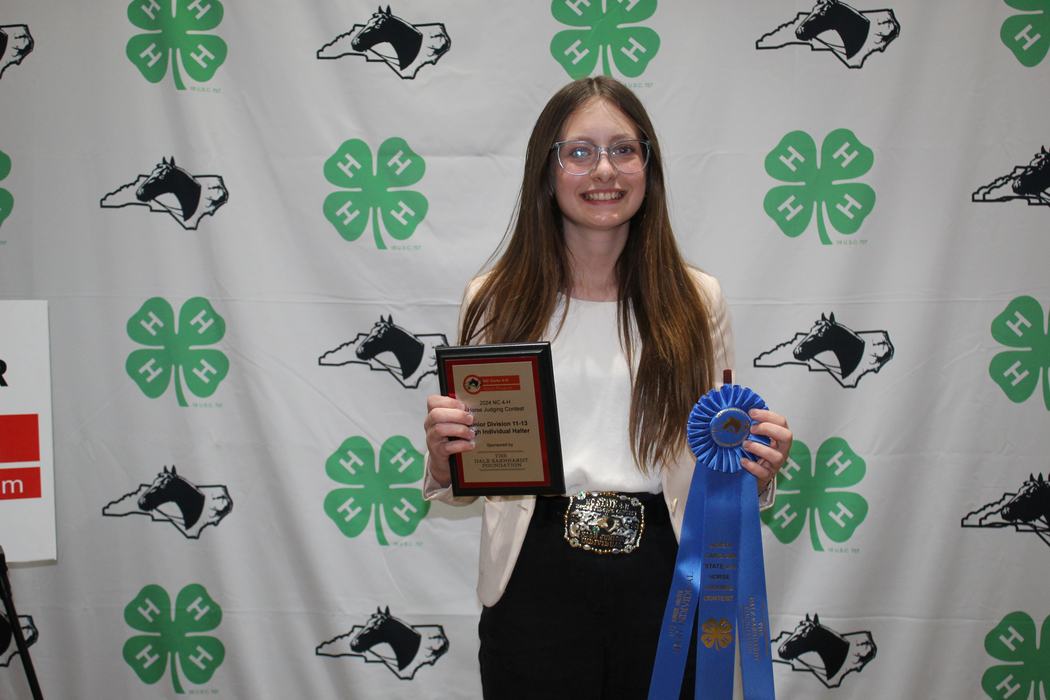 Young woman holding plaque "2024 NC 4‑H Horse Judging Contest" and large blue ribbon
